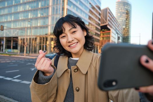 Positive asian girl takes selfie on smartphone, shows heart with fingers and makes a photo on street, tourist posing with mobile phone.