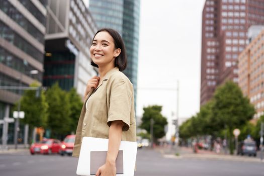 Outdoor shot of asian girl with laptop, going somewhere in city centre, walking on street, going to work.