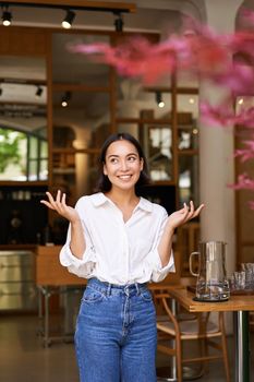 Vertical shot of enthusiastic brunette girl looking excited, raising hands up, posing near cafe entrance, inviting people to her restaurant.