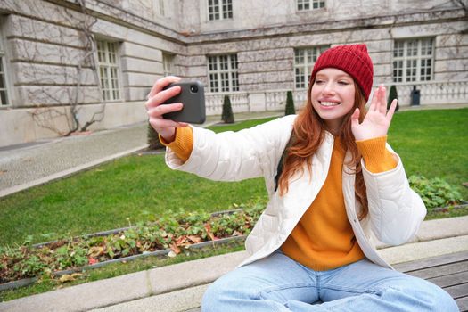 Young teen redhead girl sits on bench in park and takes selfie, makes a photo of herself with smartphone app, records vlog.