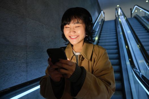 Smiling korean girl going down escalator in dark, holding mobile phone, using smartphone app, listening music, commuting in city.