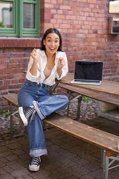 Vertical shot of enthusiastic asian girl, sits with laptop, screen is empty and blank, woman laughing and celebrating, triumphing.