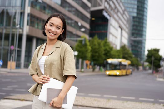 Beautiful asian girl smiles as commutes to work, stands on street with laptop and notebook.