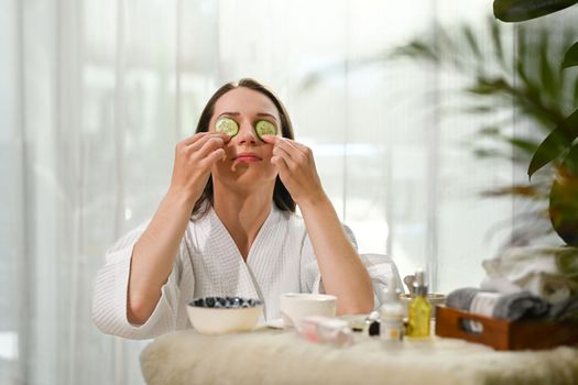 Young woman in bathrobe with cucumber slices on eyes, taking spa skincare procedures. Beauty treatment and detox concept.