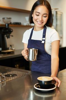 Vertical shot of smiling asian girl in cafe, giving out cappuccino, made order, prepared coffee for you, standing behind counter in blue apron.