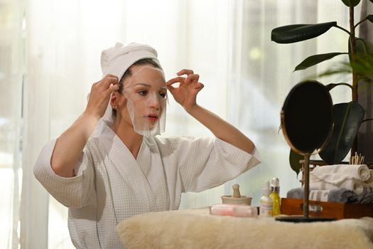 Attractive woman in bathrobe applying sheet face mask, making daily beauty routine at home. Self care concept.