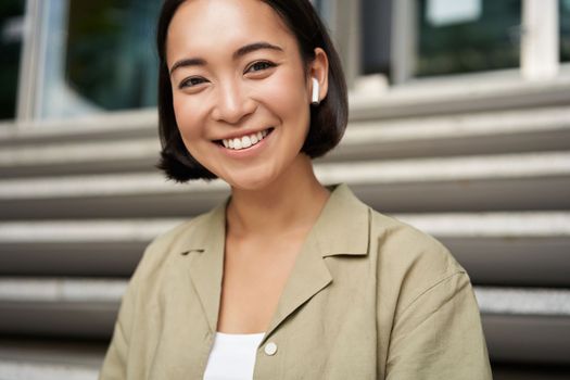 Portrait of smiling asian girl listens music, podast in wireless earphones, using headphones outdoors, sitting on street.