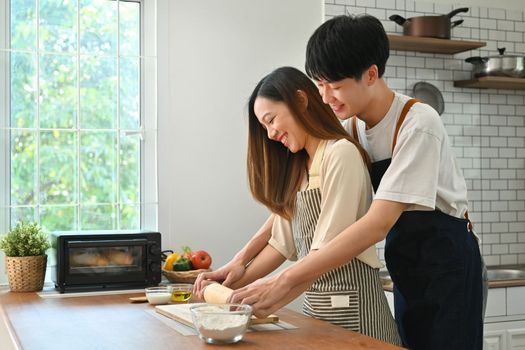 Attractive man and woman in casual outfits kneading dough with a rolling pin on wooden table in kitchen interior.