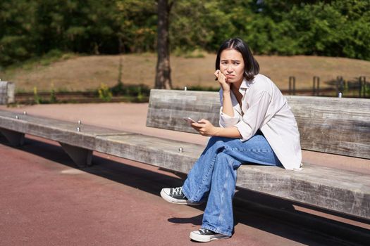 Depressed asian girl sits on bench in park with smartphone, feeling uneasy and stressed, frowning and sighing.