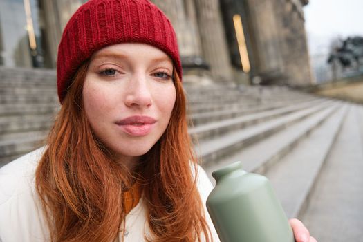 Young redhead female tourist rests during her trip, opens thermos and drinks hot tea, having a break after sightseeing.