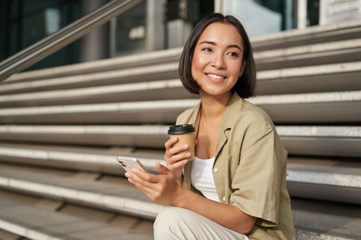 Happy urban girl drinks her takeaway coffee and scrolls feed on smartphone. Asian woman sits on stairs with tea and holding mobile phone.