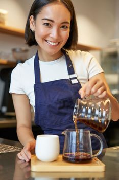 Vertical shot of smiling cute barista girl, pouring filter coffee, making order for cafe client, wearing blue apron.