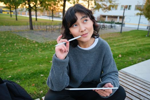 Portrait of asian woman, student in park, sits on bench with digital tablet and a pen, thinking, looking aside thoughtful, making notes, does her homework outdoors.