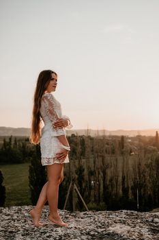 Romantic beautiful bride in white dress posing with sea and mountains in background. Stylish bride standing back on beautiful landscape of sea and mountains on sunset