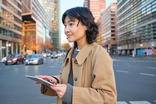 Woman walking on street using digital tablet, holding gadget in hands and looking away, standing in city centre.