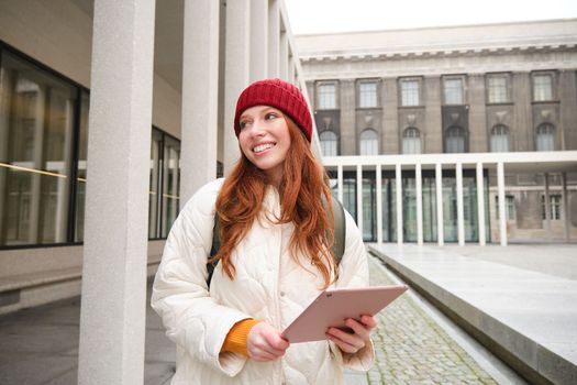 Stylish ginger girl, tourist walks with digital tablet around city, woman connects to iternet on her gadget, looking up information, texting message.