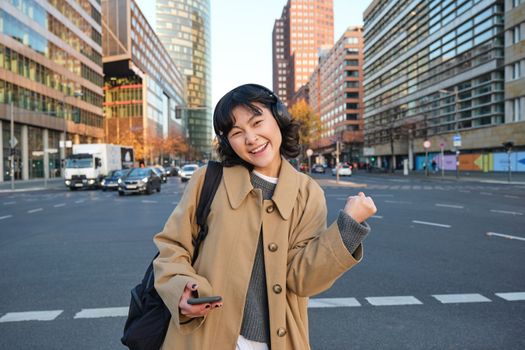 Portrait of young asian girl, student walks in city, listens music in headphones and uses mobile phone on streets, celebrates, triumphs and makes joyful fist pump.