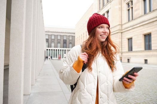 Beautiful smiling girl, tourist with backpack, holding smartphone, using map on mobile phone application, looking for sightseeing in internet app, standing outdoors.