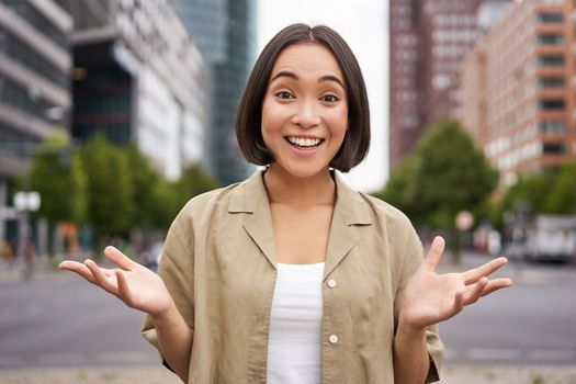 Modern asian girl standing on street with surprised face, looking impressed and amazed, hear awesome news.