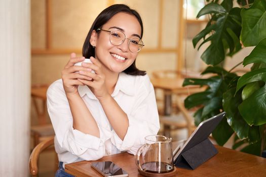 Smiling asian girl in glasses, woman working on remote, drinking coffee and using tablet. Copy space