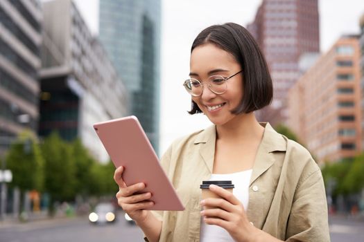 Woman standing on street, drinking coffee, reading on tablet, checking messages, using app.