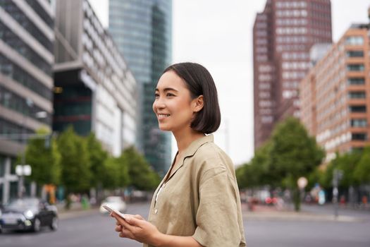 Young asian woman exploring city with smartphone app, holding mobile phone and walking on screet.