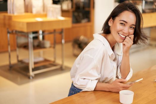 Portrait of young asian woman, sitting in cafe, holding smartphone, chatting and messaging while drinking coffee.