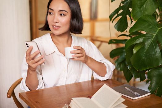 Young asian businesswoman sitting in cafe with notebook, coffee and looking at smartphone, video chat, calling someone.