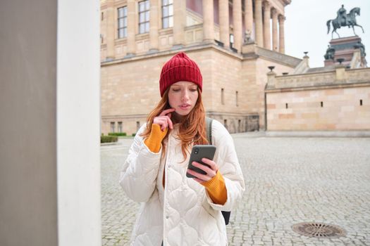 Smiling redhead girl tourist, walks around city and explores popular landmarks, sightseeing, holding smartphone, looking at her mobile app and checks with city map.