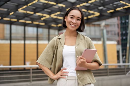 City girl standing with tablet on street, using application, smiling at camera.