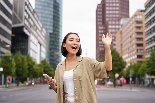 Happy asian girl passing by friend and waving at them on street, saying hello while walking in city, holding smartphone.