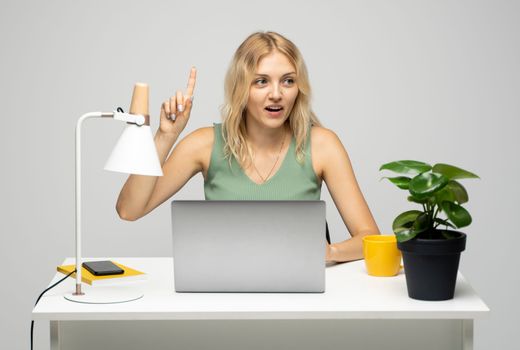 Overjoyed happy blonde woman pointing finger up and smiling toothy while sitting at a table with a laptop. Having idea, business plan, startup concept