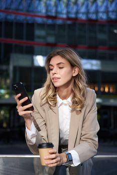 Vertical shot of businesswoman drinks coffee, looks at mobile phone app. Corporate woman on her lunch break, using smartphone.