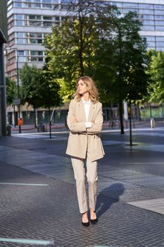 Portrait of young confident corporate woman in beige suit, standing on street in her business outfit, looking self-assured.