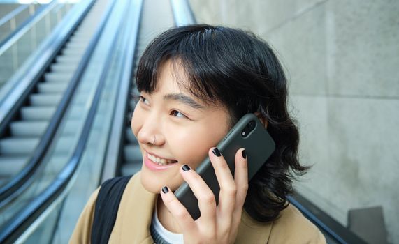 Headshot of smiling korean woman with smartphone, makes a phone call, goes down escalator in city, commutes to university.