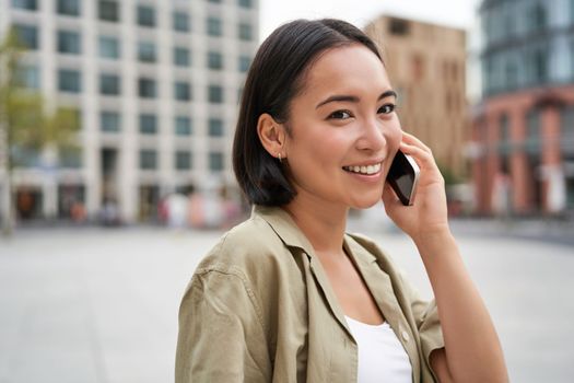 Cellular connection. Young asian woman makes a telephone call, talking on mobile smartphone and walking on street.