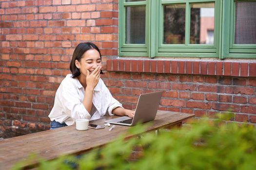 Stylish young urban girl in outdoor cafe, sitting on bench with laptop, smiling, browing on computer.