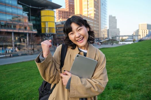 Happy korean girl, student celebrates, makes fist pump and triumphs, holds tablet and smiles pleased, winning, standing on street.