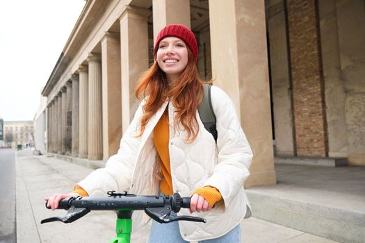 Young smiling redhead girl, student rides electric scooter, rents it and travels around city. Transport and travelling concept