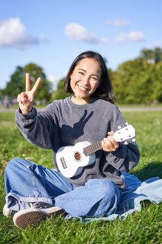 Vertical shot of positive asian girl shows peace sign, plays ukulele in park, rests and enjoys the day. Copy space.