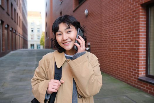 Cellular technology concept. Young modern girl talks on mobile phone, walks on street and smiles, has conversation over telephone. Copy space