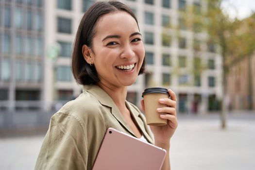 Portrait of asian girl with tablet, drinks coffee on street, walking in city centre.