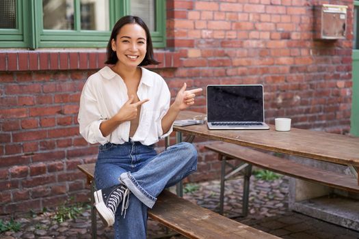 Stylish young asian woman sitting in cafe with laptop, outdoor table, pointing finger at advertisement, showing banner copy space.