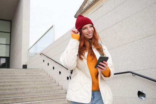 Happy girl student in red hat, holds smartphone, tourist looks at map app on her phone, explores sightseeing, texts message, looks for couchsurfing, rents place to stay online.