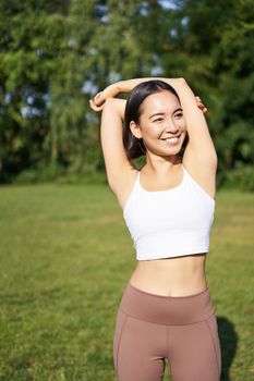 Asian woman stretching her arms, doing fitness workout in park, smiling pleased, warming up before jogging on fresh air in daytime.