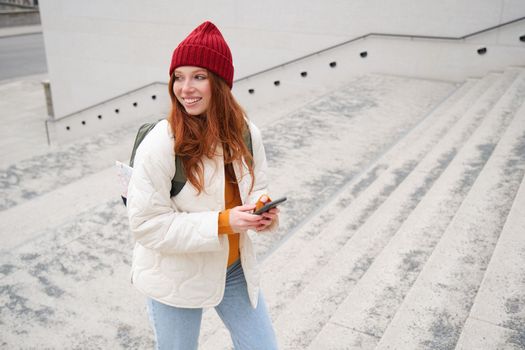 Joyful girl tourist looks at mobile phone, texts message on smartphone social media application, walks around city, looks for sightseeing on mobile app.