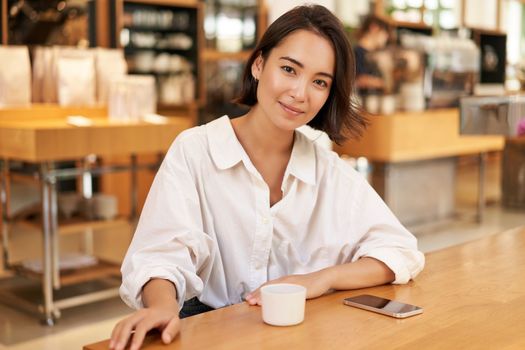 Portrait of confident asian woman, sitting in cafe, smartphone and coffee on table. Businesswoman smiling with confidence. Copy space