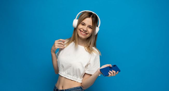 Portrait of beautiful blonde young woman in white headphones with a smartphone listening to music on blue background