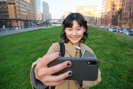 Happy young woman, student takes selfie on street, holds her tablet and homework notes, uses smartphone. Copy space