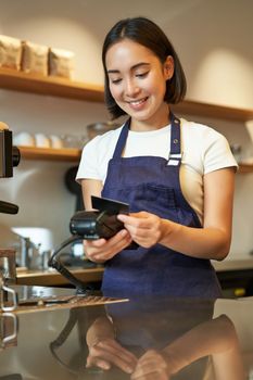 Beautiful smiling asian girl, barista insert client credit card in POS terminal, processing payment, taking orders in coffee shop.